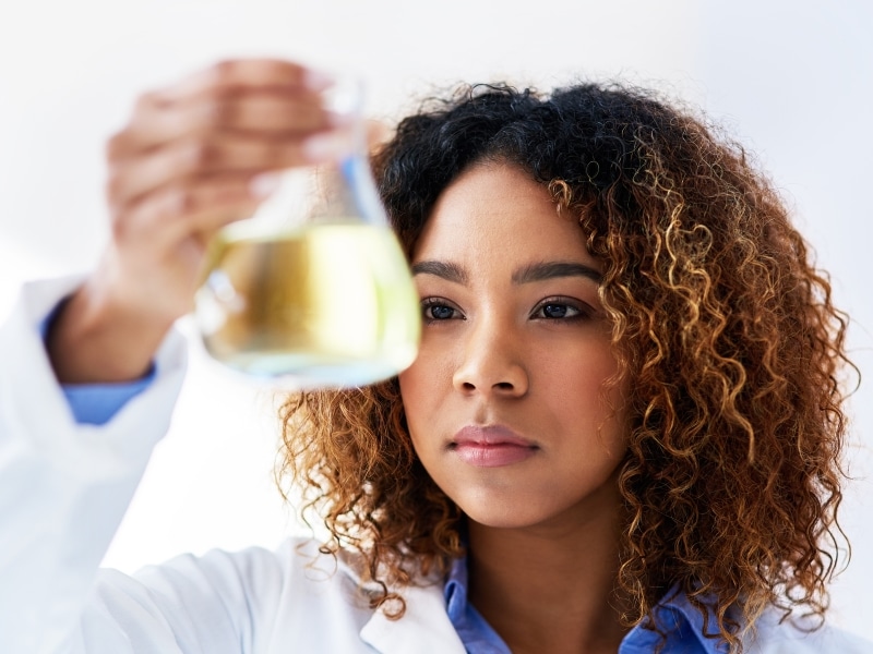 A female scientist with dark hair, wearing a white lab coat, inspecting the quality of a flask of oil for My Swim Cream.